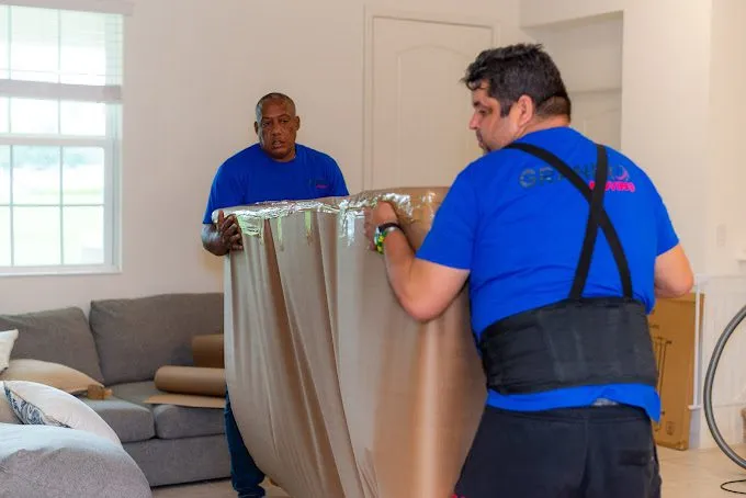Two Granero Moving movers in blue company t-shirts wrapping a sofa in protective paper